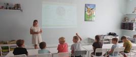 Kobieta w kremowej sukience stoi zwrócona w stronę grupy dzieci siedzących na białych krzesłach. Chłopiec w jasnych włosach podnosi rękę do góry. Za kobietą na ścianie znajduje się wyświetlona grafika z zaleceniami dotyczącymi zdrowego żywienia. 