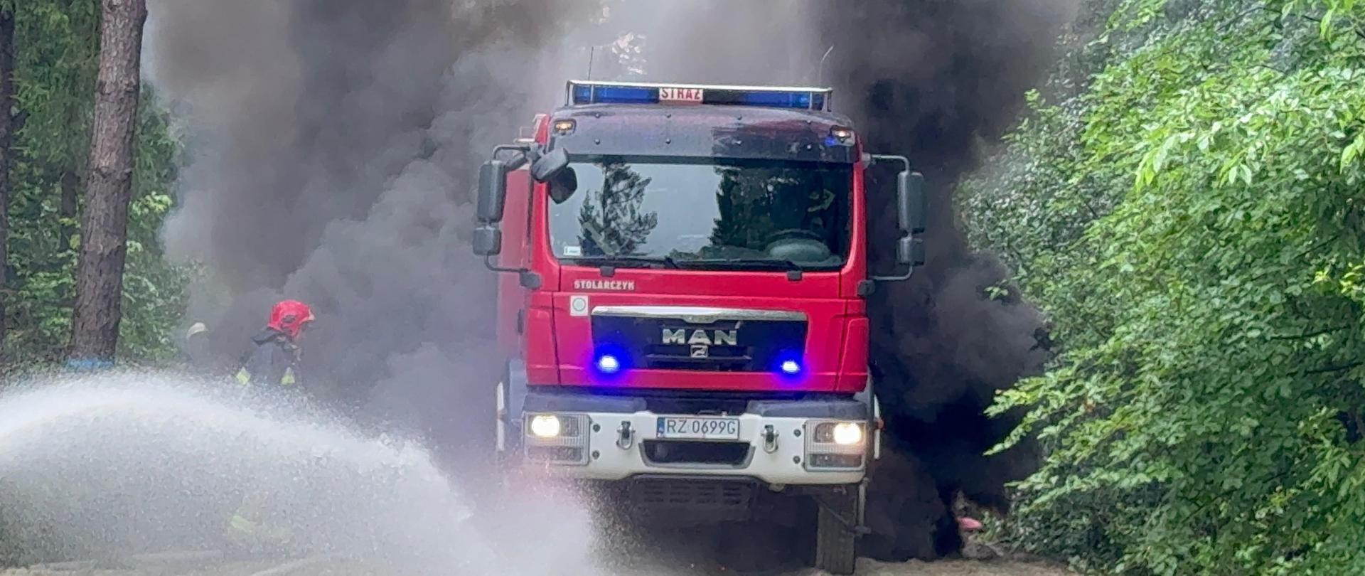 Fotografia wykonana podczas wojewódzkich ćwiczeń ratowniczo-gaśniczych "Mielec 2024". Na zdjęciu gaszenie pożaru samochodu gaśniczego w lesie. Na pierwszym planie widać strażaka, który podaje prąd wody na palący się pożar. W tle widać kłęby czarnego dymu symulujące pożar samochodu. Samochód stoi na drodze leśnej z włączonymi światłami sygnałowymi.