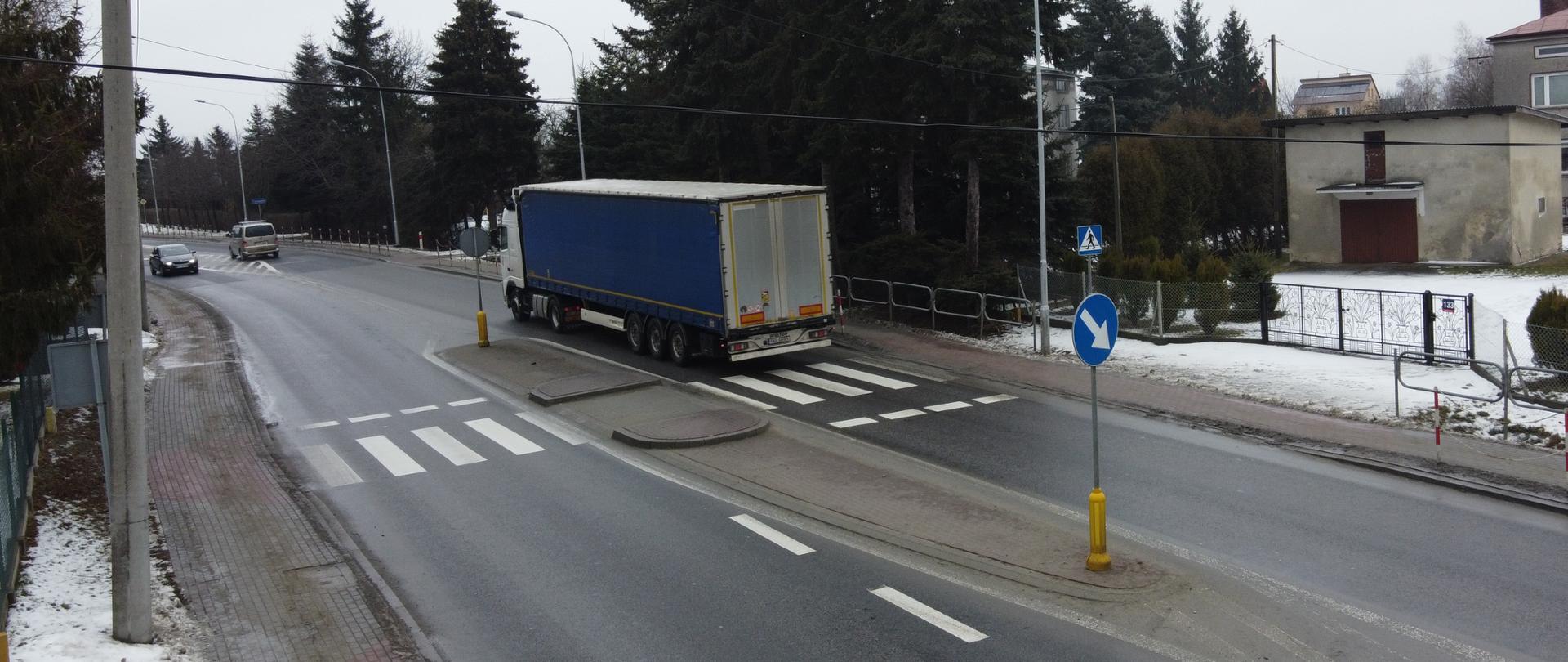 Pochmurno, zima. Widok na drogę. Jezdnia czarna, na poboczach leży śnieg. Widok na przejście dla pieszych. Auta jadą w obu kierunkach.