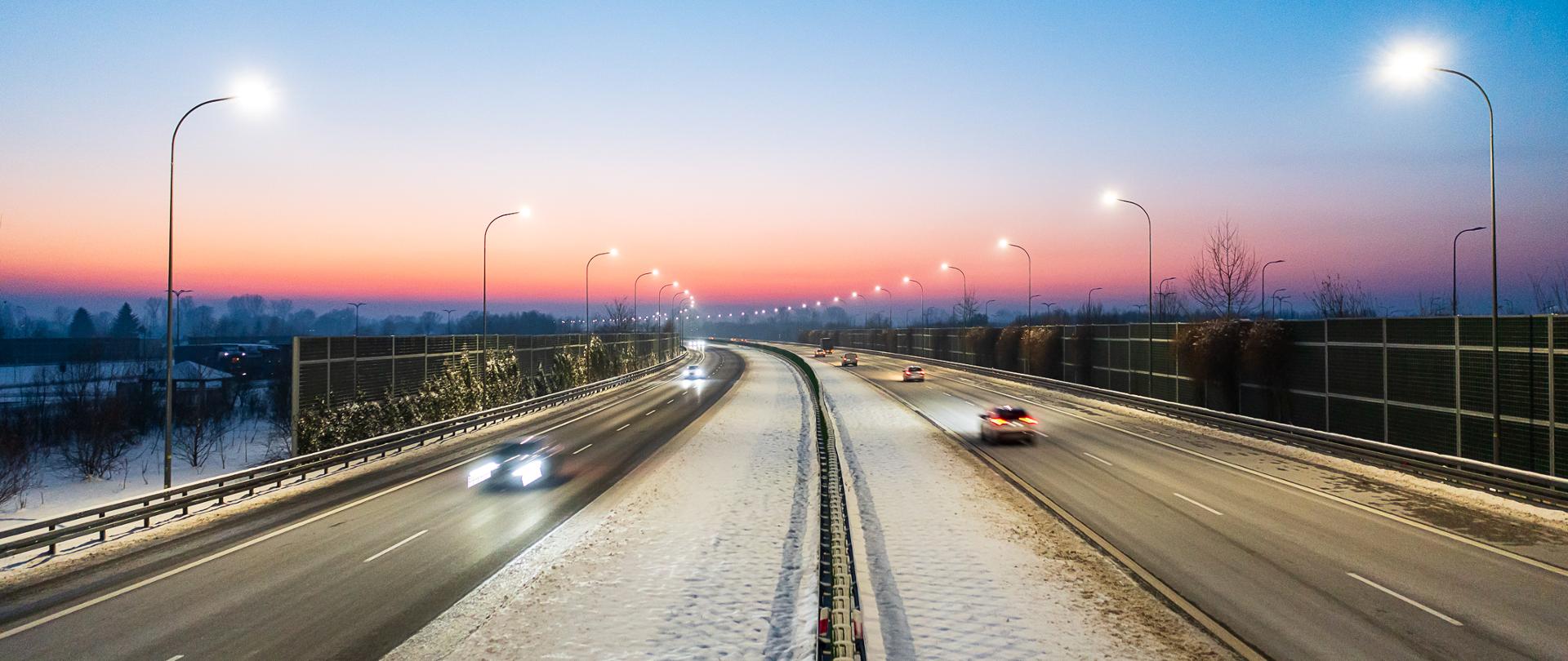 Widok z poziomu ziemi na autostradę A4. Zima, pora dnia zaraz po zachodzie słońca. Niebo w kolorach różowo - fioletowych. Na jezdni widać jadące auta. Na pasie rozdziału i poboczu leży śnieg. Po obu stronach drogi świecą latarnie. 