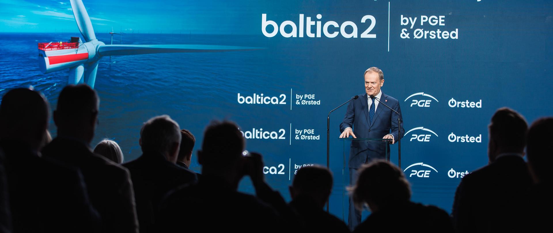 Na zdjęciu widać oficjalne wydarzenie związane z rozpoczęciem budowy morskiej farmy wiatrowej Baltica 2. Na scenie stoi mężczyzna (Premier RP Donald Tusk) w garniturze, przemawiający przy mównicy. W tle znajduje się duży ekran wyświetlający grafikę turbiny wiatrowej na morzu oraz tekst informujący o projekcie. Publiczność obserwuje wydarzenie.