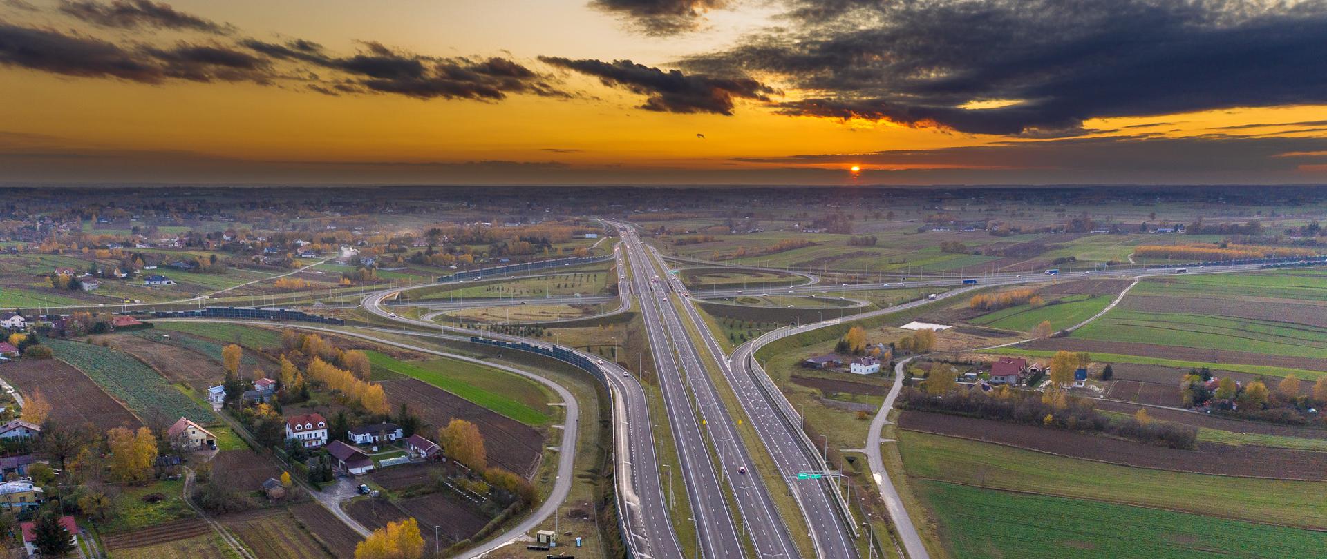 Zdjęcie lotnicze drogi węzła Lublin Sławinek na połączeniu dróg ekspresowych S12 i S17. Zdjęcie wykonane przy zachodzie Słońca. Widoczny ruch pojazdów na obu drogach ekspresowych. W tle widoczne zabudowania oraz pola uprawne i łąki.