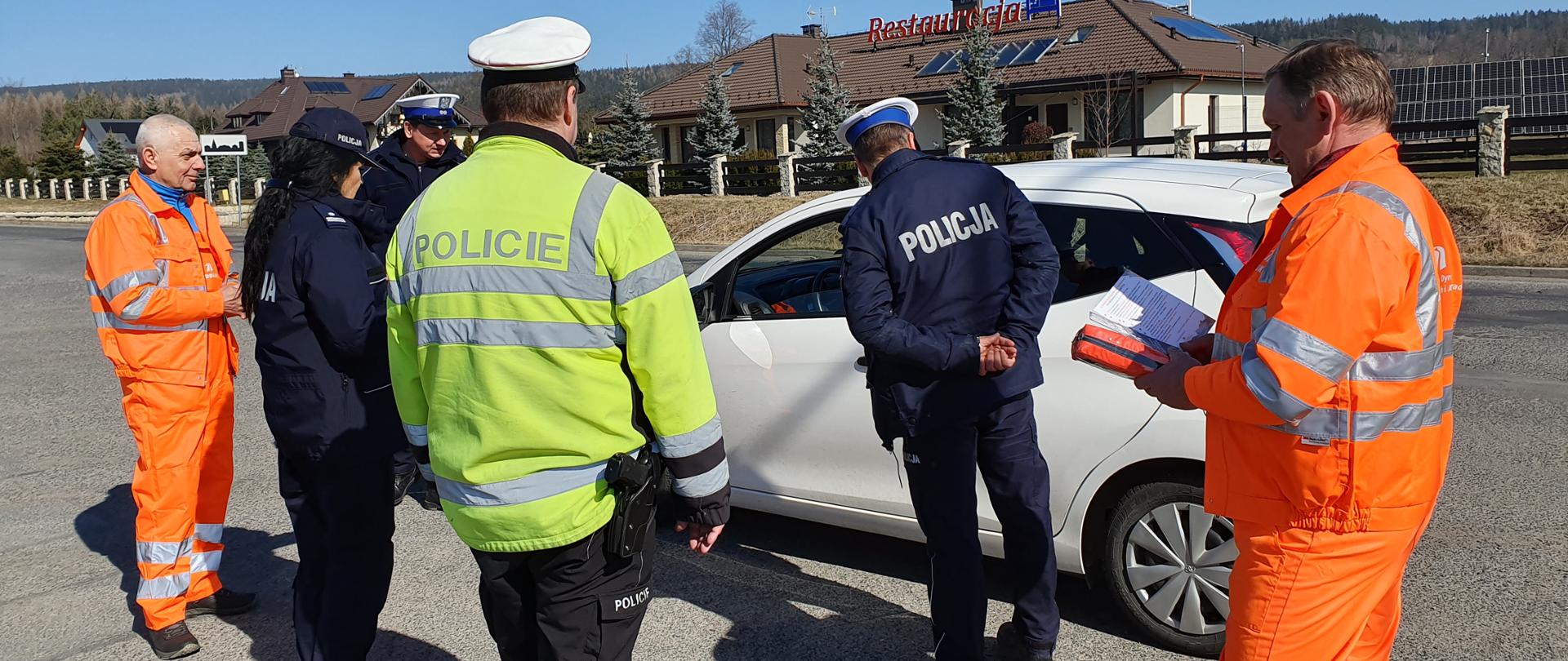 na zdjęciu widoczni czterej policjanci i dwóch pracowników GDDKiA stojących przy zatrzymanym samochodzie. W ręku pracownika GDDKiA apteczka samochodowa i ulotka GDDKiA