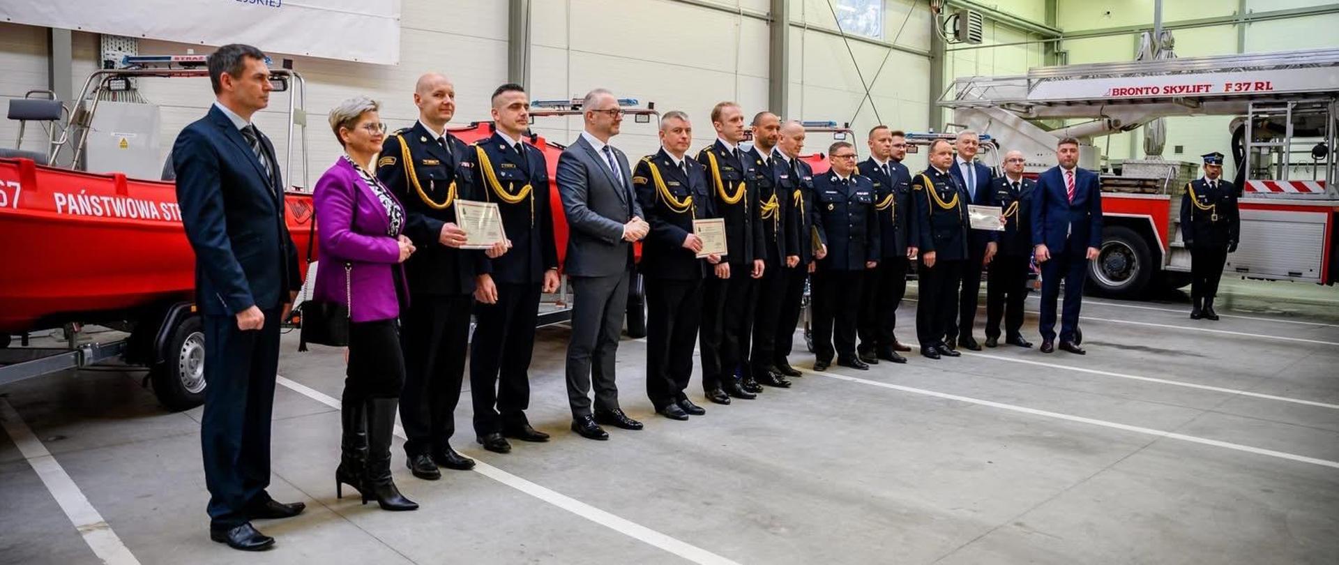 Funkcjonariusze Państwowej Straży Pożarnej i druhowie Ochotniczych Straży Pożarnych stoją w hali garażowej. W tle widoczne pojazdy pożarnicze.