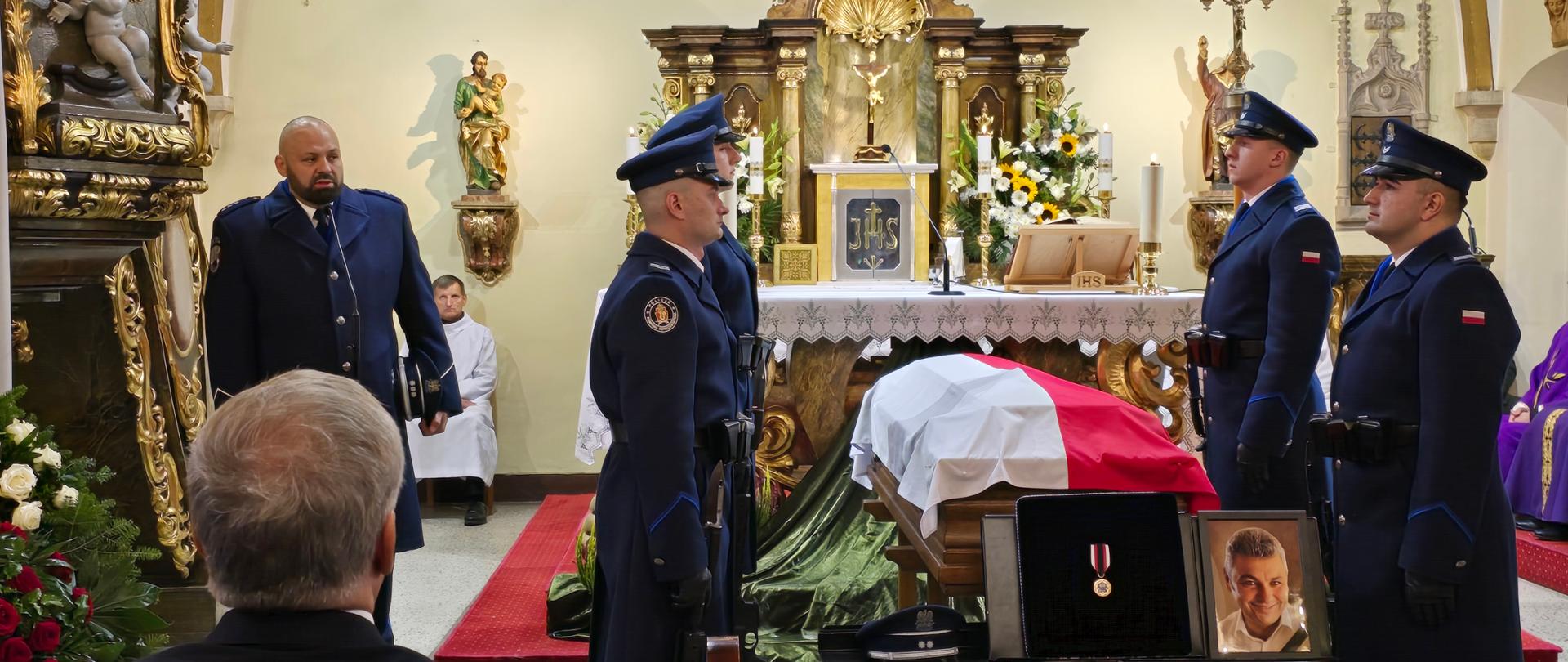 Ujęcie z wnętrza kościoła. Trumna przykryta biało-czerwoną flagą. Obok stoją policjanci w mundurach galowych.