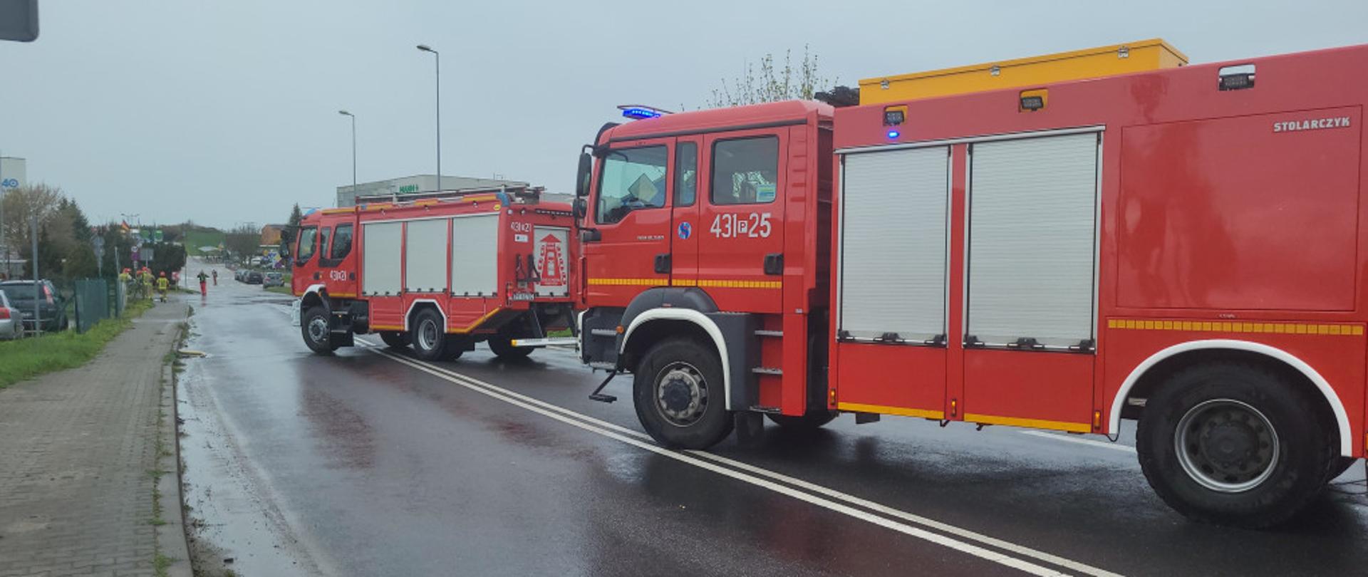 Zdjęcie przedstawia 2 wozy strażackie stojące na jezdni, podczas zdarzenia związanego z wyciekiem substancji żrącej na jezdnię. Samochody mają włączone sygnały świetlne w postaci niebieskich świateł błyskowych.