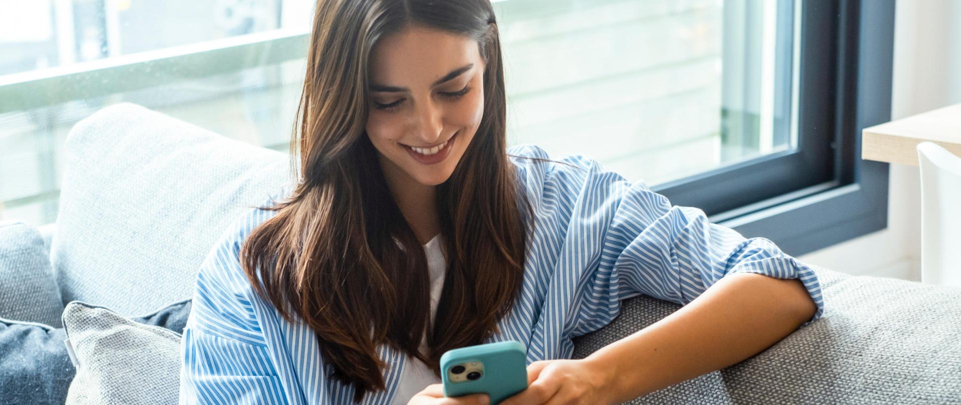 Happy girl checking social media while holding smartphone at home. Smiling young brunette woman using mobile phone app to play games, shop online, order delivery while relaxing on sofa. Scrolling.