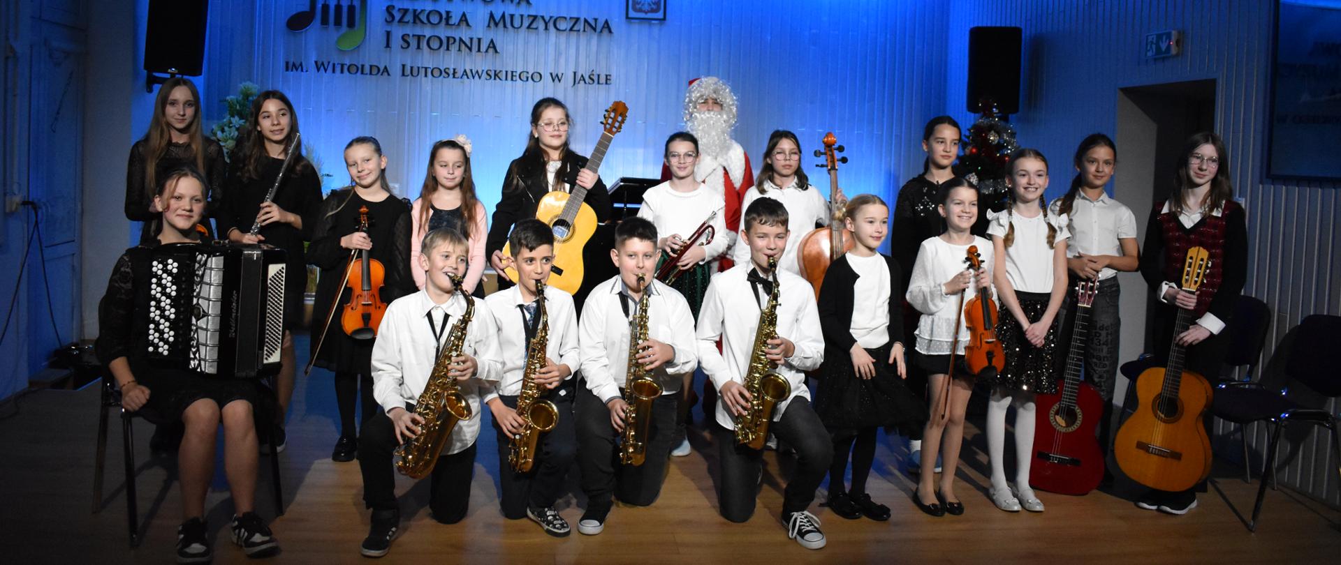 Na zdjęciu znajduje się grupa uczniów z instrumentami na scenie w auli PSM, w tle na ścianie logo szkoły i godło państwowe, kolorystyka niebieska