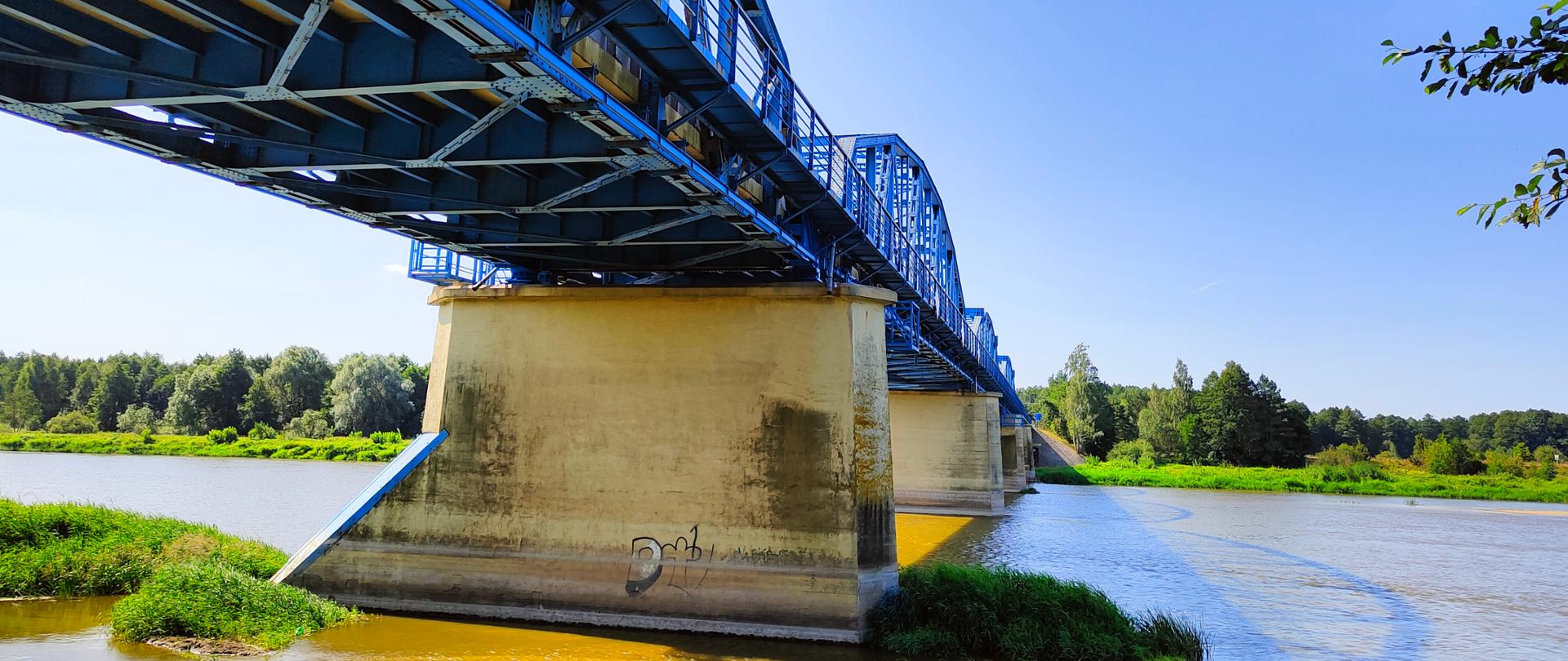 Zdjęcie z boku mostu, widok na filary posadowione w rzece i spód mostu pomalowany na niebiesko