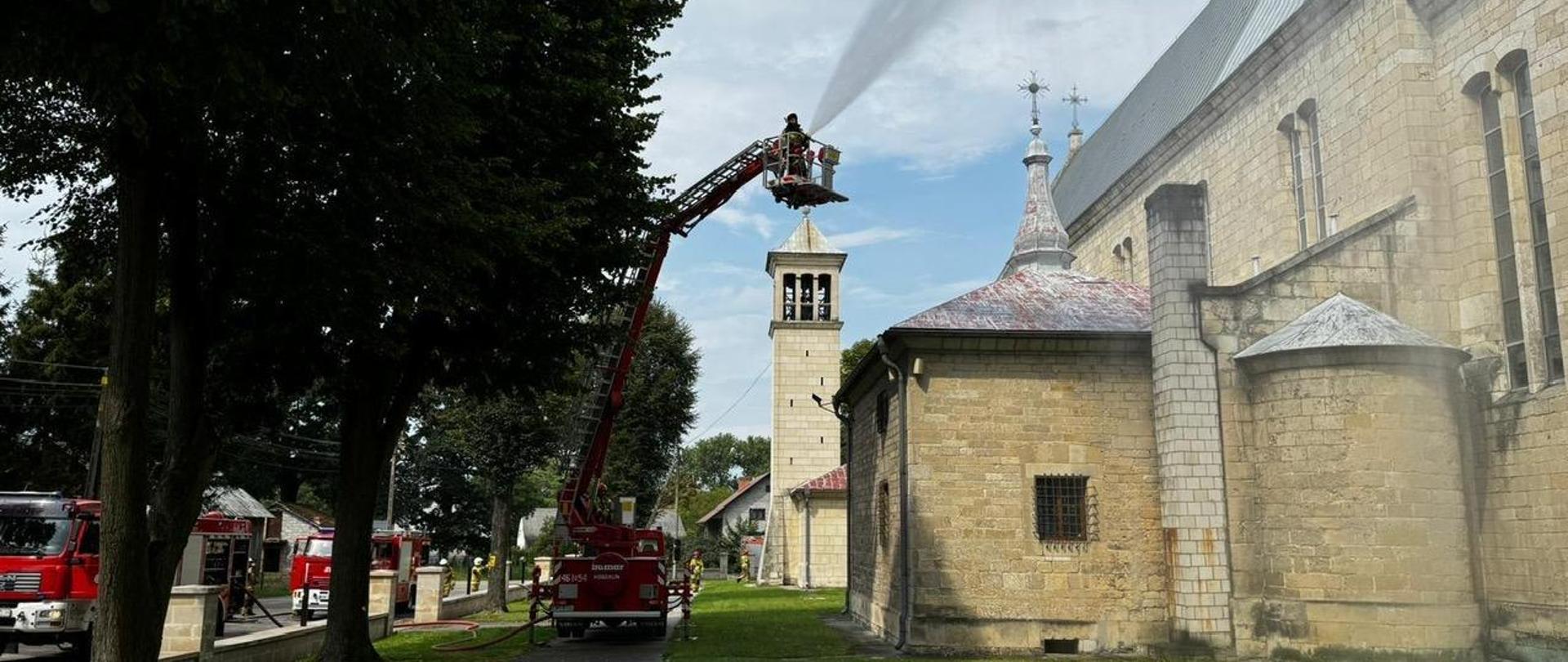 Na zdjęciu widoczny podnośnik strażacki w koszu strażak podający prąd wody z działka na dach kościoła