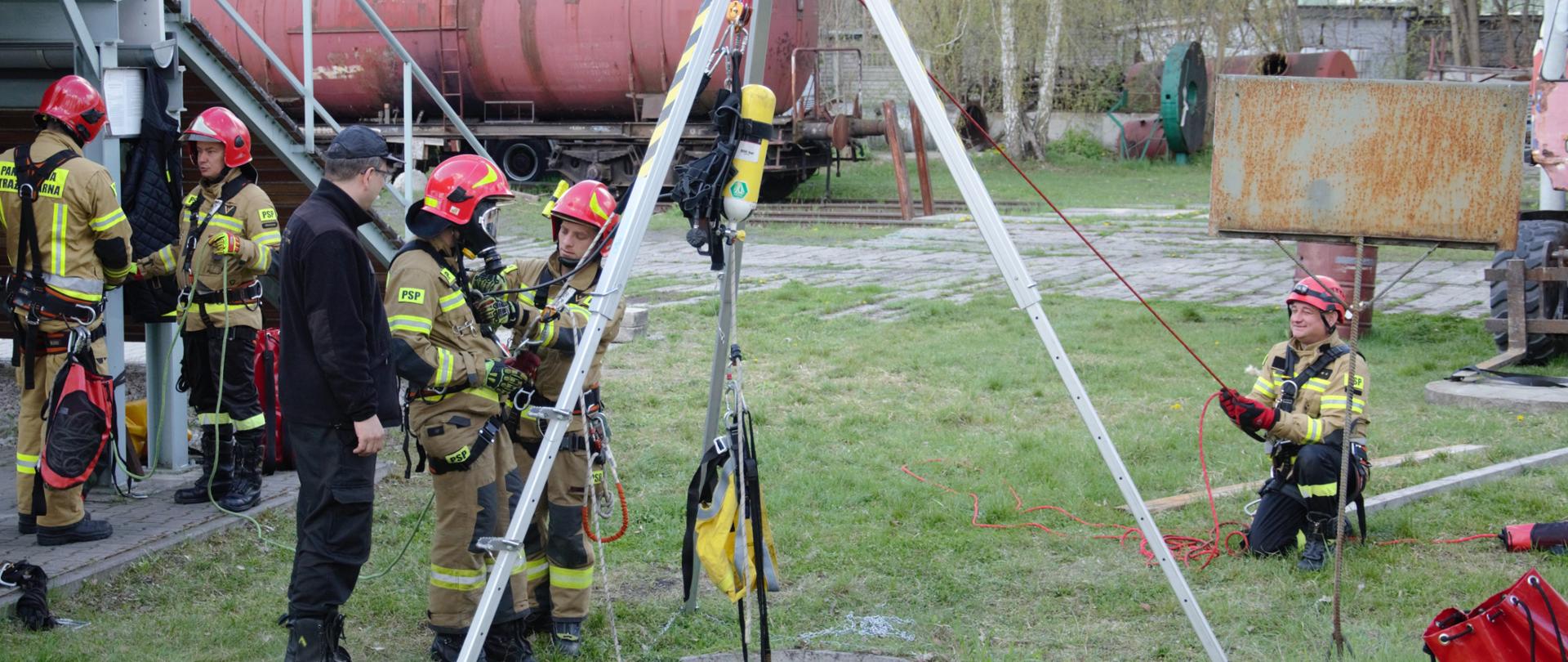 Strażacy ćwiczą wyciąganie poszkodowanego ze studni na trójnogu. ubrani sa w ubranie specjalne strażackie