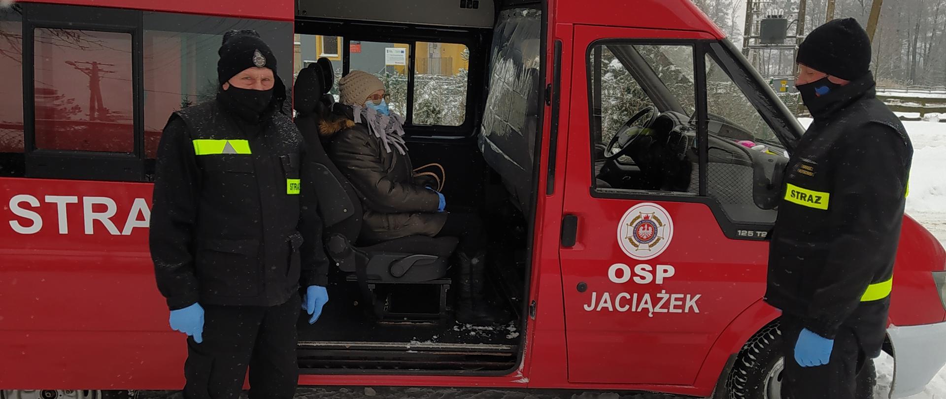 Dwóch strażaków OSP stoi przed samochodem strażackim. W środku, w tylnym rzędzie siedzi starsza kobieta transportowana do przychodni POZ. Drzwi do samochodu są otwarte.