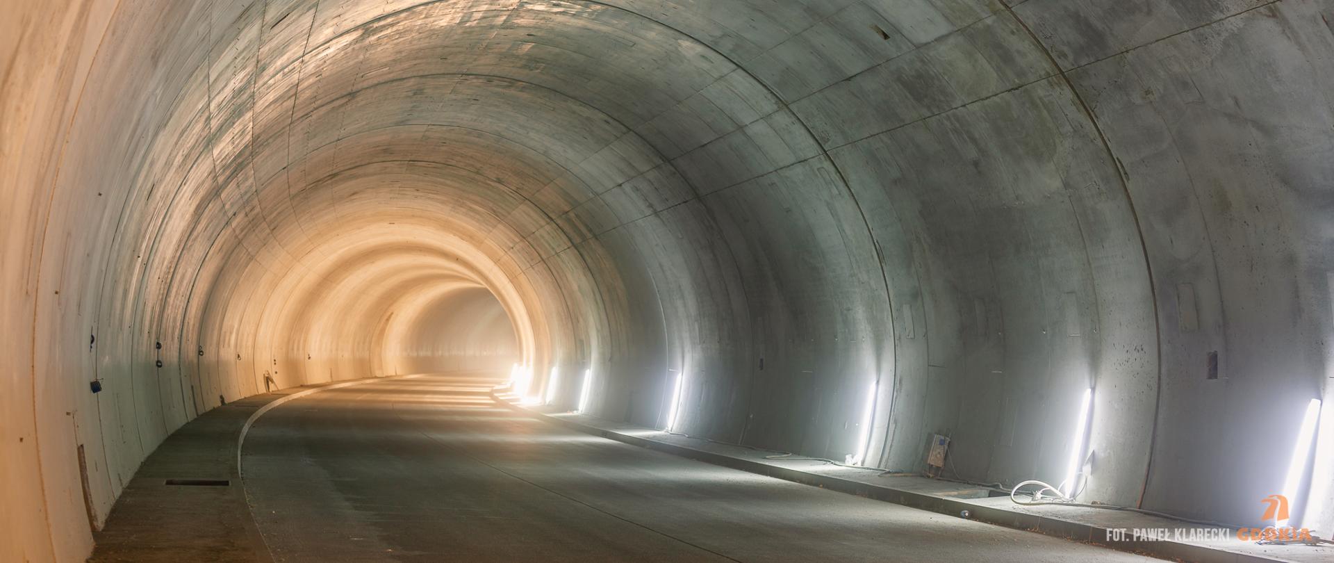 Zdjęcie przedstawia wnętrze tunelu drogowego. Tunel jest oświetlony jasnym światłem, które tworzy efektowne refleksy na jego betonowych ścianach. Ściany tunelu są gładkie i mają lekko zaokrąglony kształt. Na podłodze tunelu widać asfaltową nawierzchnię drogi. W tunelu nie ma żadnych pojazdów ani ludzi, co sprawia, że wygląda on na pusty i spokojny. 