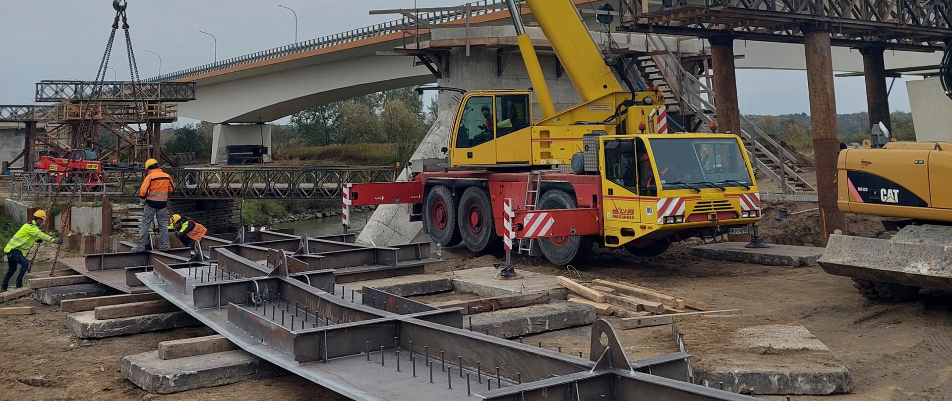 Budowa mostu w Sandomierzu - trzech robotników w kaskach i kolorowych ubraniach ochronnych przygotowuje długi płaski szary stalowy element do przeniesienia przez dźwig. Trwa montaż zaczepów dźwigowych. Z tyłu żółto-czerwony dźwig. W tle drugi most nad rzeką