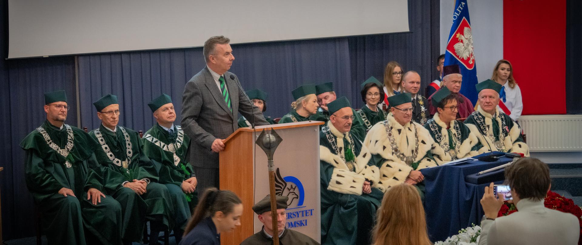 Minister Wieczorek stoi za mównicą, mówi do mikrofonu, za nim w dwóch rzędach krzeseł siedzą ludzie w zielonych ceremonialnych strojach.