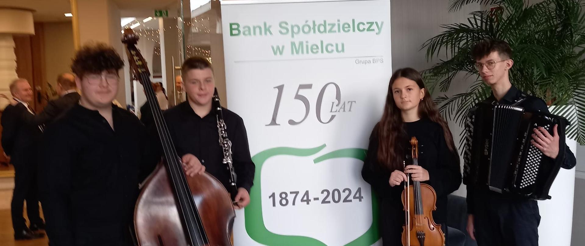 Na zdjęciu widoczna jest grupa czterech młodych muzyków, stojących przed roll-upem z napisem „Bank Spółdzielczy w Mielcu – 150 lat 1874-2024”. Osoby trzymają instrumenty: kontrabas, klarnet, skrzypce i akordeon. W tle znajduje się eleganckie wnętrze z elementami roślinnymi i lampami sufitowymi.