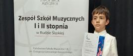 Chłopiec stoi trzymając dyplom w rękach, z anim baner Zespołu Szkół muzycznych I i II st. w Rudzie Śląskiej