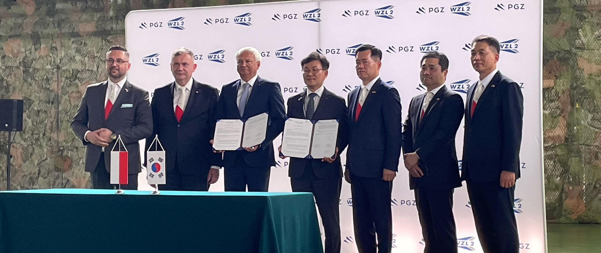 Za stołem stoją przedstawiciele strony polskiej oraz koreańskiej. Dwóch mężczyzn trzyma w dłoniach dokumenty. Na stole stoją proporczyki - polski i południowokoreański. W tle ścianka z logotypami PGZ oraz WZL 2. 