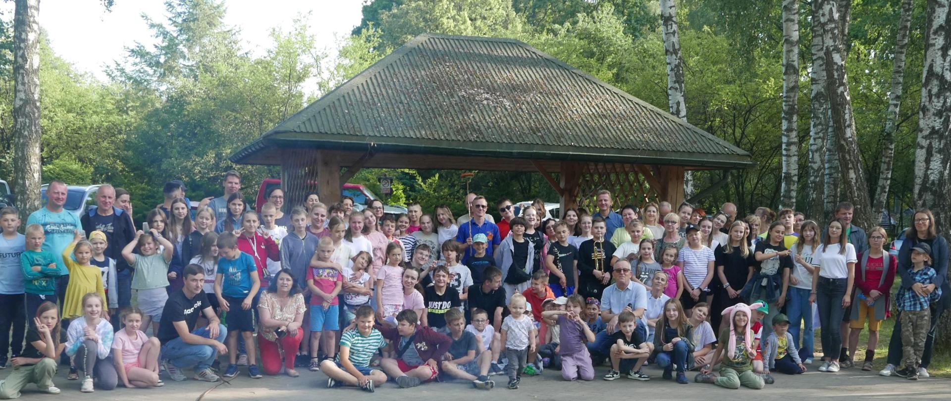 Zdjęcie grupowe z lasem w tle. Dużo dzieci, młodzieży i dorosłych na zdjęciu. Kolorowe koszulki i uśmiechy. Dalej widać wiatę ze spadzistym dachem i zieleń lasu.