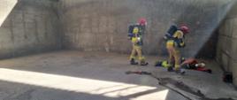 Zdjęcie przedstawia dwóch strażaków w umundurowaniu specjalnym z założonymi aparatami ochrony dróg oddechowych. Strażacy przeszukują halę magazynową paliw alternatywnych. Koło nich, na ziemi leży poszkodowana osoba. Strażacy przystępują do działań.