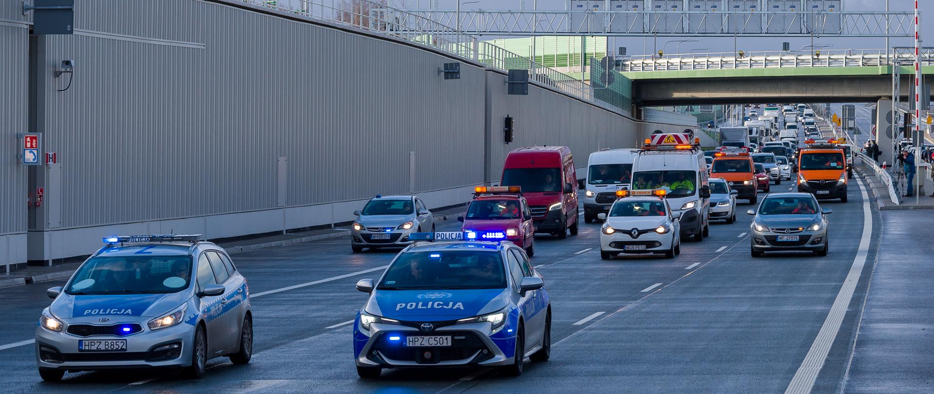 
Zdjęcie przedstawia scenę na wielopasmowej drodze z wieloma pojazdami, w tym dwoma radiowozami z migającymi niebieskimi światłami, czerwonym vanem oraz kilkoma innymi samochodami i pojazdami serwisowymi GDDKiA. Wszystkie pojazdy poruszają się za dwoma radiowozami na pierwszym planie.