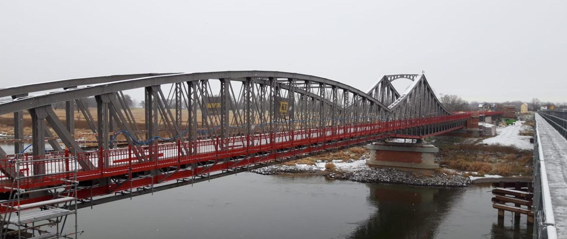 Stalowy most z barierkami w kolorze czerwonym z widocznymi elementami rusztowania związanego z trwającym remontem. Poniżej przepływa Ścinawa. Na moście, brzegu i w tle niewielka warstwa śniegu po niedawnych opadach.