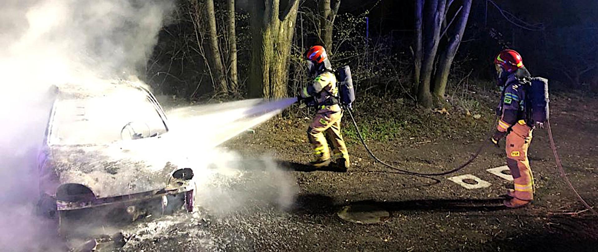 Zdjęcie przedstawia dwóch strażaków w ubraniach specjalnych i aparatach ochrony układu oddechowego gaszących pianą gaśniczą płonący pojazd.