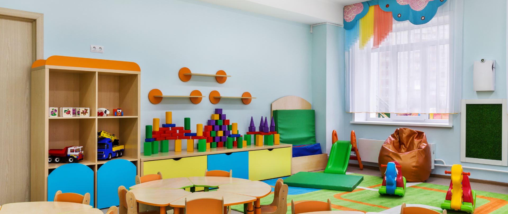 Widok sali przedszkolnej: na pierwszym planie okrągłe stoliki i krzesełka z prawej strony zabawki dziecięce. Za stolikami meble z zabawkami dla dzieci. Wszystko w żywych kolorach.