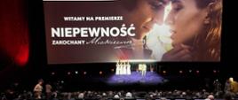 Widok z tyłu na salę kinową i ekran. W fotelach siedzą ludzie, na scenie kilka osób ubranych na biało, na ekranie wyświetlany jest zwiastun filmu z tekstem: Witamy na premierze „Niepewność. Zakochany Mickiewicz”