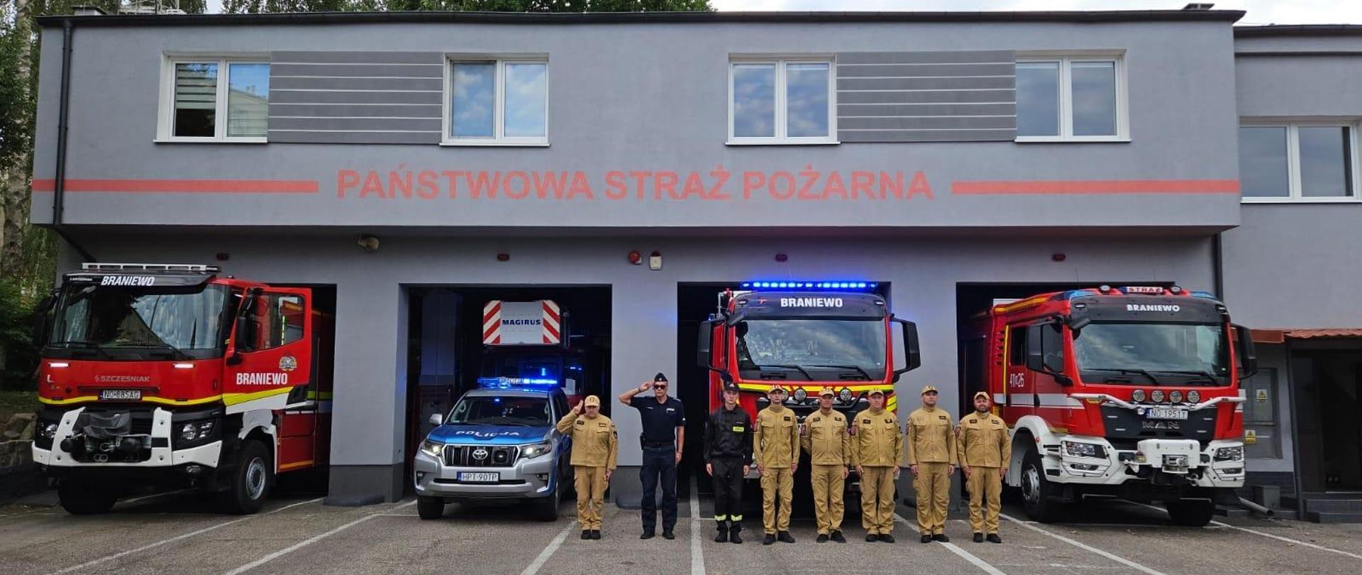 Strażacy w mundurach i policjant ustawieni w jednym szeregu na tle czterech samochodów ratowniczych i remizy jednostki ratowniczo gaśniczej.