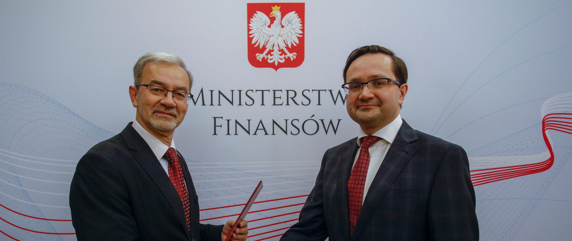 Minister Jerzy Kwieciński wraz rzecznikiem finansowym Mariuszem Goleckim na tle banera Ministerstwa Finansów