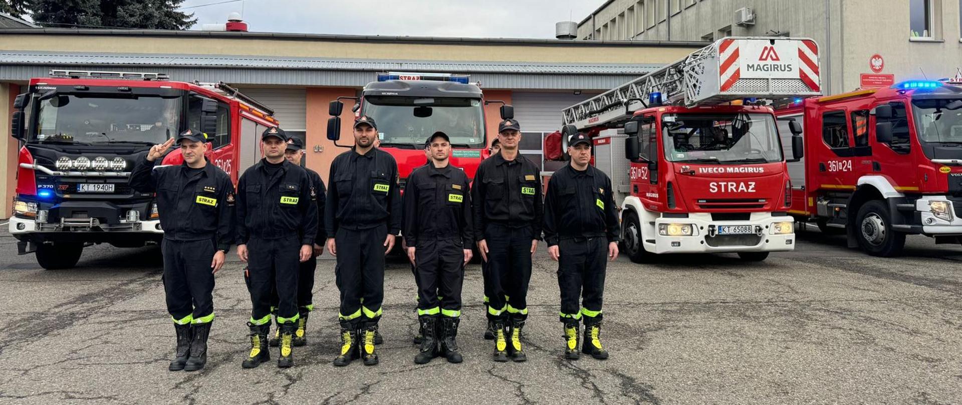 Na uroczystej zbiórce przed budynkiem remizy stoją w dwuszeregu strażacy ubrani w umundurowanie koszarowe koloru czarnego. Za strażakami stoją ustawione obok siebie samochody pożarnicze w tle budynek Jednostki Ratowniczo Gaśniczej