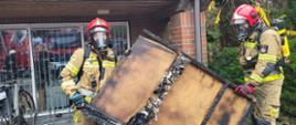 Na zdjęciu widzimy 2 strażaków wynoszących nadpalone elementy konstrukcyjne na zewnątrz budynku.