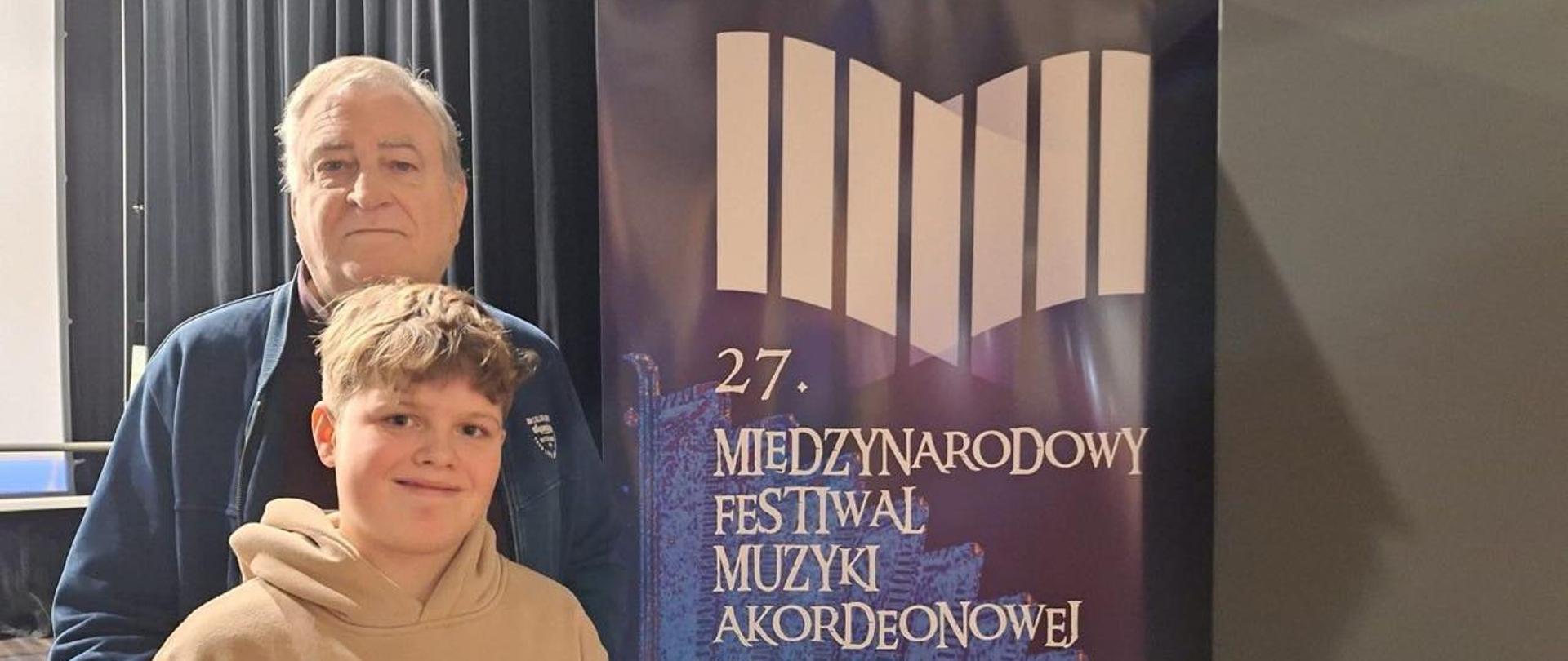 Mikołaj Piątek oraz nauczyciel Zdzisława Szymczyka na zdjęciu przy banerze reklamującym 27. Międzynarodowy Festiwal Muzyki Akordeonowej w Przemyślu