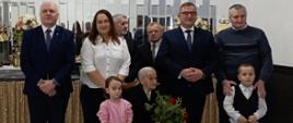 Jubilatka siedzi na krześle stojącym w uroczyście udekorowanej sali. Obok niej, po lewej stoi mała dziewczynka, po prawej kilkuletni chłopiec. z tyłu stoją kobieta i czterech mężczyzn. Przed wszystkimi stoją dwa duże białe kosze z kolorowymi bukietami kwiatów.