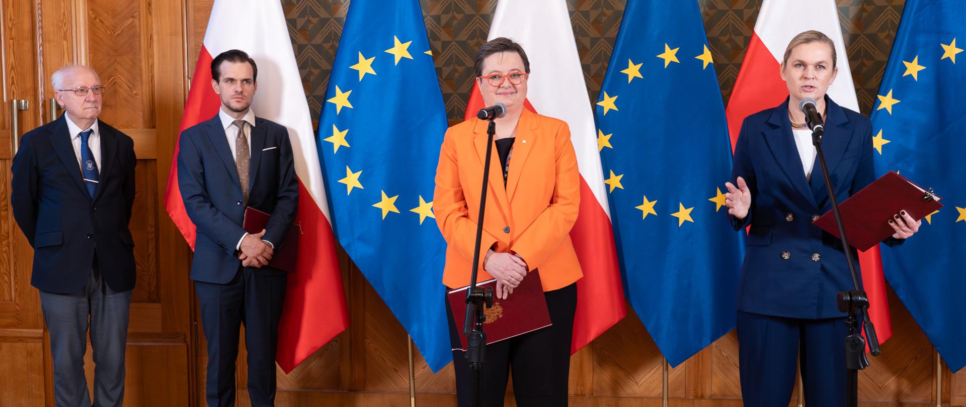 Duża sala ze ścianami w zielony wzorek, Minister Nowacka stoi przy mikrofonie i przemawia obok niej wiceminister Katarzyna Lubnauer, po lewej stronie stoi jeszcze dwóch mężczyzn. W tle flagi Polski i UE