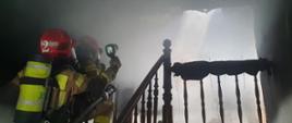 Na zdjęciu dwóch strażaków na zadymionej klatce schodowej. Jeden ze strażaków wykonuje pomiary trzymaną w ręce kamerą termowizyjną