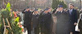 Czterech strażaków w mundurach wyjściowych salutuje przed grobem generała Tadeusza Buka. Za nimi stoją żołnierze i osoby cywilne. Przed nimi ustawione są wieńce z kwiatami.
W tle widać las. 