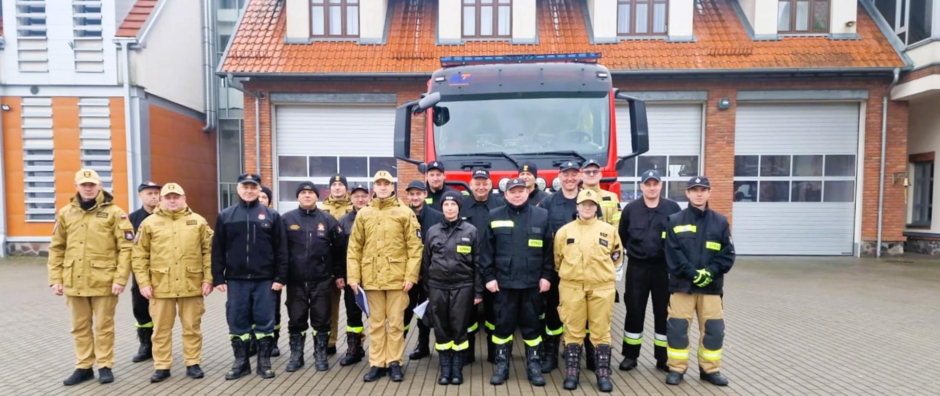 Zdjęcie przedstawia uczestników szkolenia podstawowego strażaka ratownika Ochotniczych Straży Pożarnych, realizowanego w Komedzie Powiatowej Państwowej Straży Pożarnej w Gołdapi wspólnie z komisją egzaminacyjną i zmianą służbową, na tle pojazdu pożarniczego zlokalizowanego przed garażami strażnicy