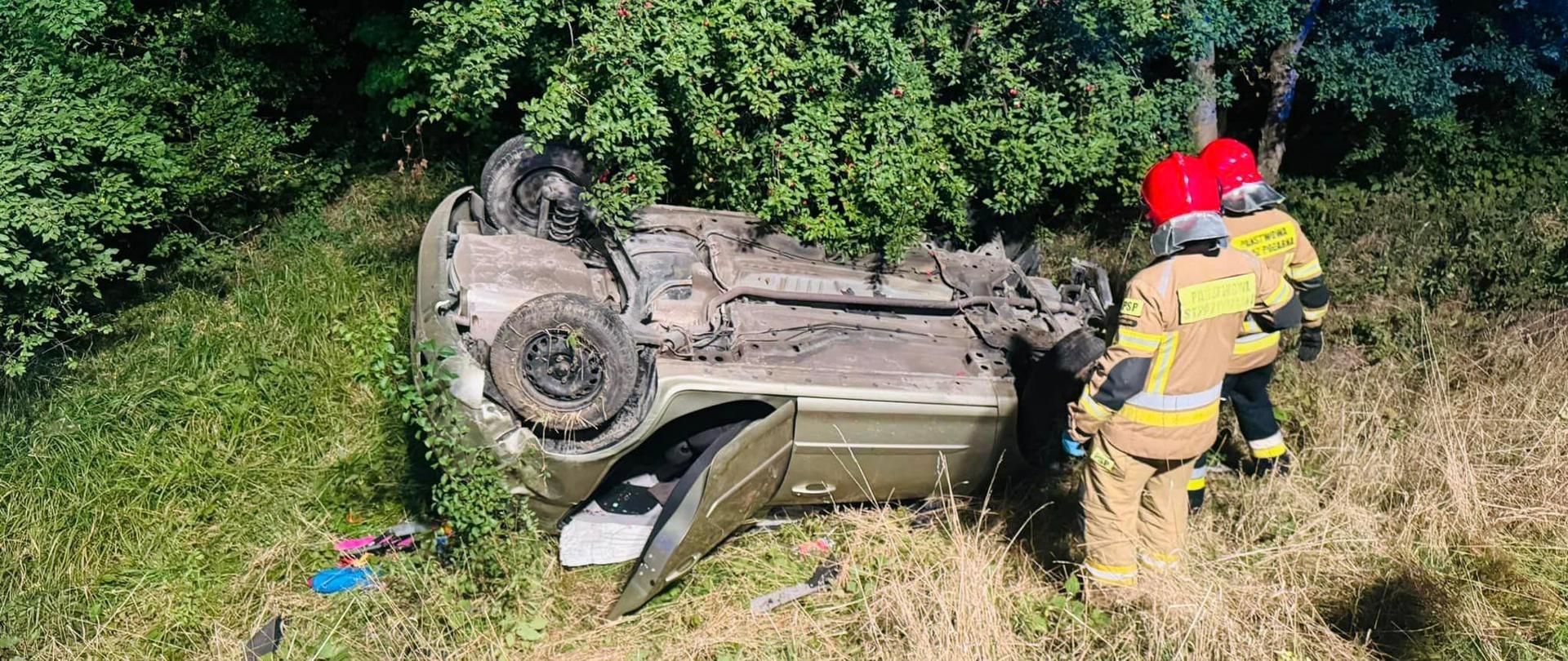 Na zdjęciu znajduje się samochód po uprzednim dachowaniu oraz pracujący przy nim strażacy.