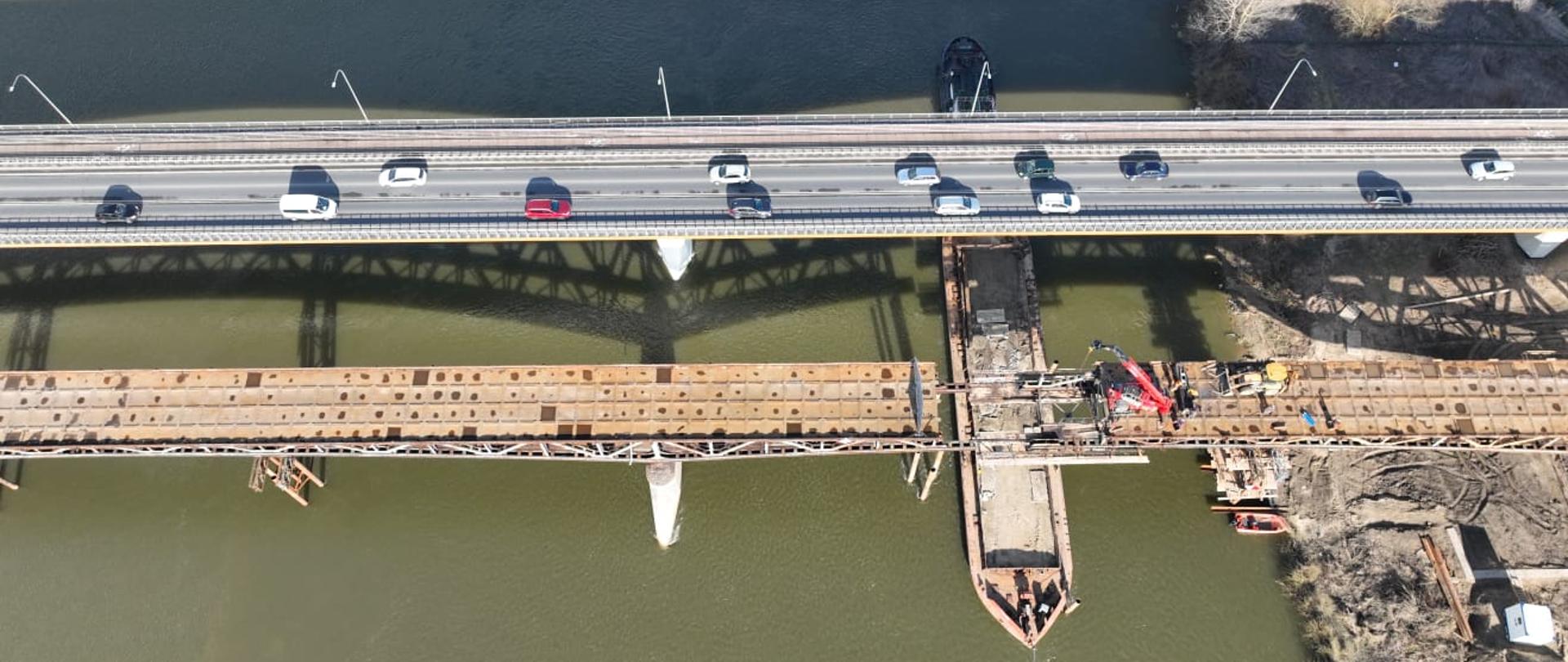 DK77 mosty w Sandomierzu - dwa mosty sfotografowane z drona. Jednym odbywa się ruch w obu kierunkach - widoczne samochody. Drugi równoległy jest rozbierany. W miejscu rozcięcia konstrukcji stalowej pod mostem zacumowana barka. Na konstrukcji dźwig i ładowarka.
