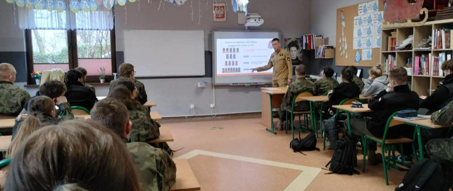 Zdjęcie wykonane w sali klasowej. Na pierwszym planie uczniowie klasy wojskowej ubrani w zielone moro. Na drugi planie, strażak w mundurze piaskowym stojący przy tablicy na której włączona jest prezentacja.