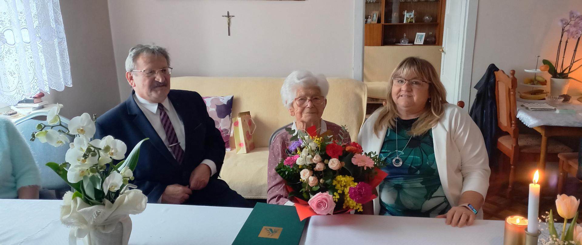Jubilatka z bukietem kwiatów siedzi przy stole, po lewej siedzi Kierownik Wydziału Świadczeń Wojciech Zielonka, po prawej Kierownik Placówki Elżbieta Akierman