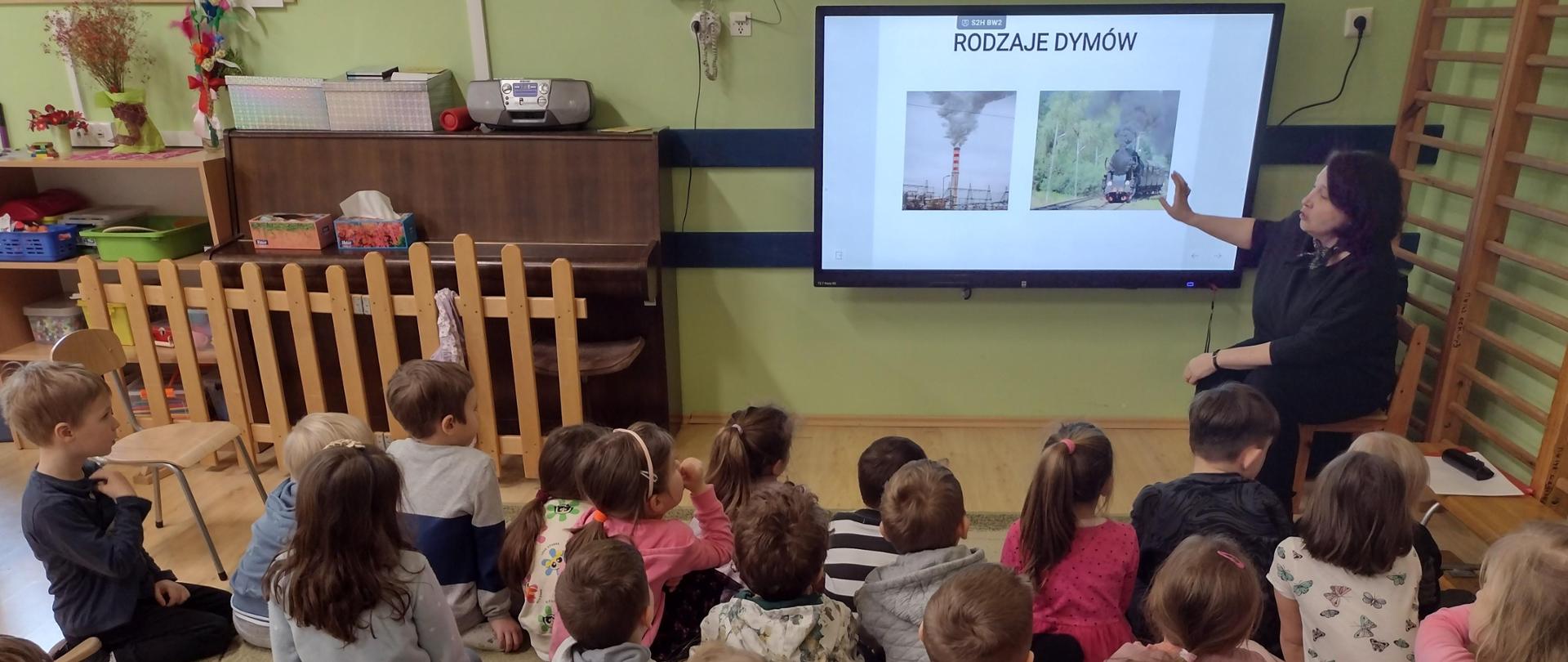 Dzieci siedzą na dywanie i oglądają prezentację z rodzajami dymów
