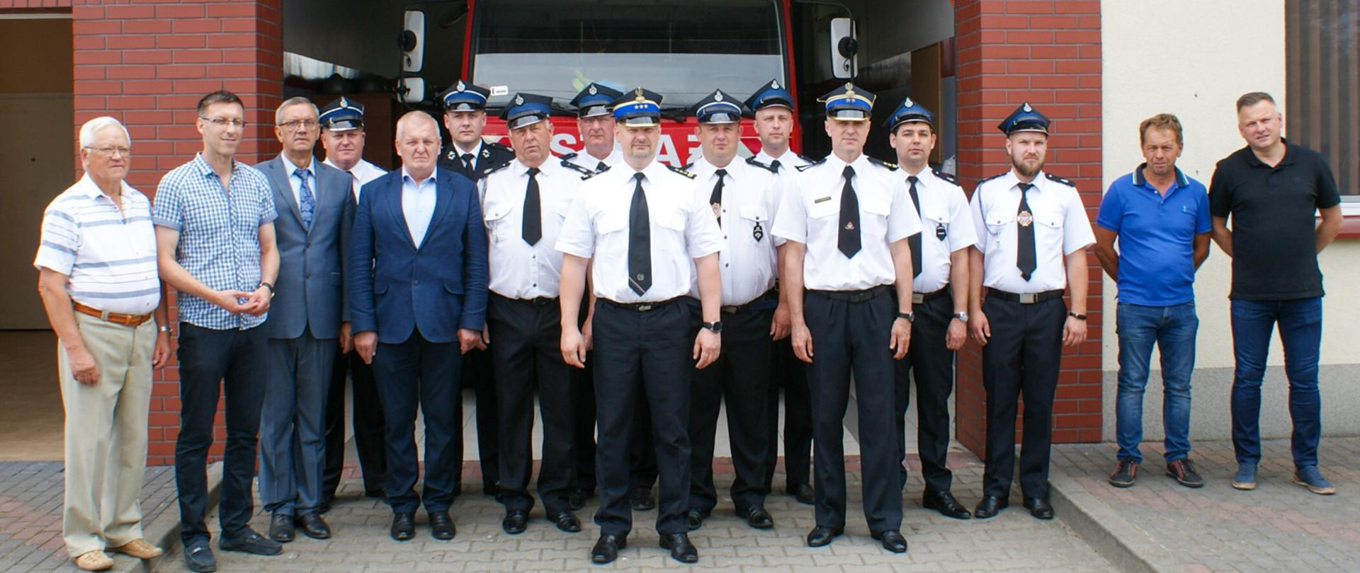 Zdjęcie pamiątkowe uczestników spotkania w OSP w Zamościu na tle strażnicy i samochodu gaśniczego w garażu.