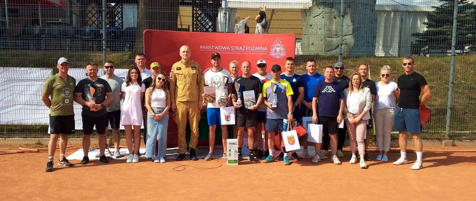 Zdjęcie zrobione na korcie tenisowym, pomarańczowa mączka. Na zdjęciu grupa zawodników z nagrodami oraz organizatorzy turnieju. Słonecznie.