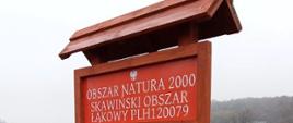 Drewniana tablica z daszkiem. Na czerwonym tle znajduje się biały napis - Obszar Natura 2000 Skawiński Obszar Łąkowy PLH120079