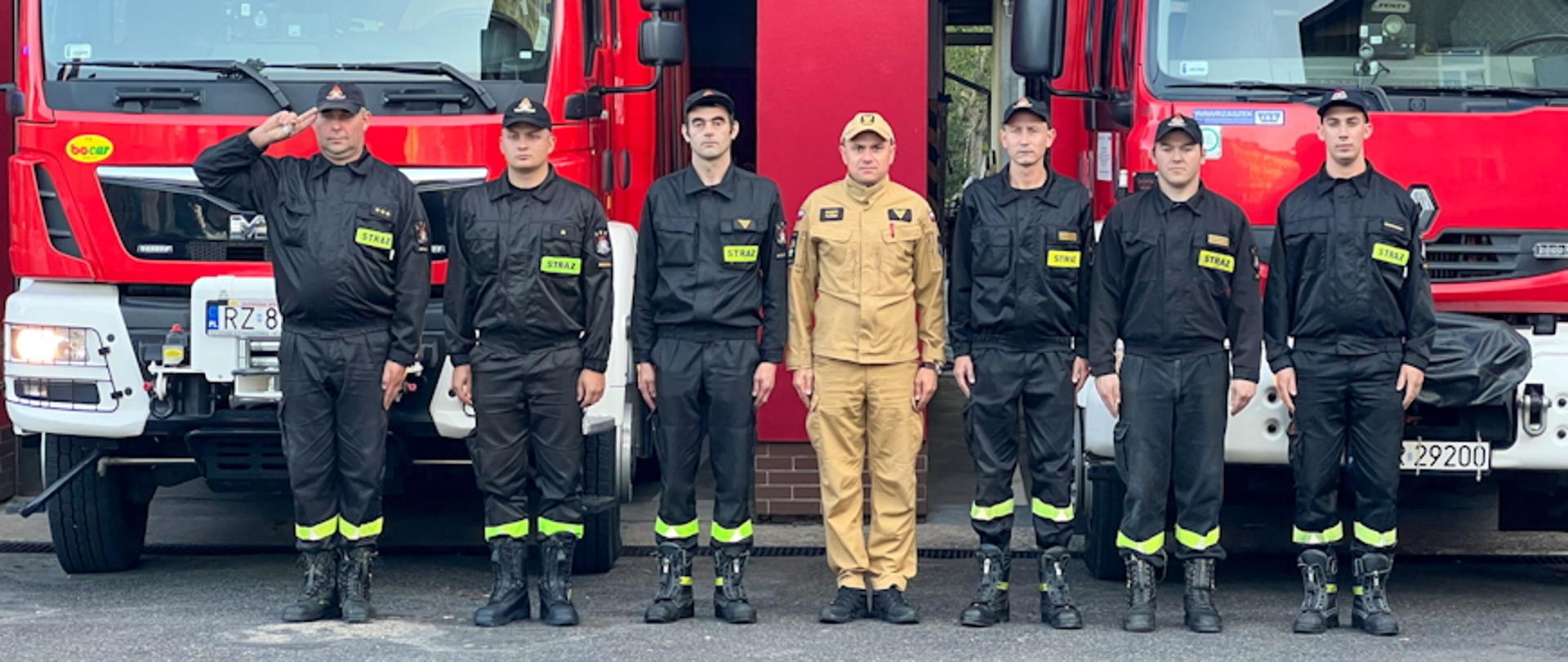 Kolorowa fotografia wykonana w pochmurny dzień na zewnątrz. Przedstawia strażaków stojących w szeregu z dowódcą oddającym honor na tle samochodów pożarniczych z włączonymi światłami dźwiękowymi i świetlnymi.
