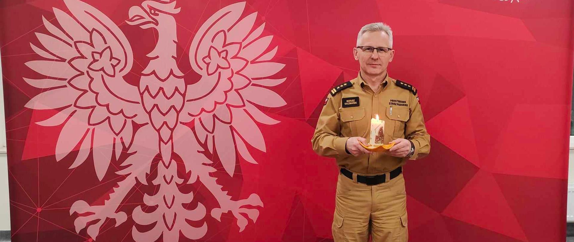 Komendant Powiatowy Państwowej Straży Pożarnej w Brzezinach starszy brygadier Mariusz Kaczmarski na tle ścinki reklamowej Państwowej Straży Pożarnej w ręku trzyma świecę z przekazanym Betlejemskim Światłem Pokoju.