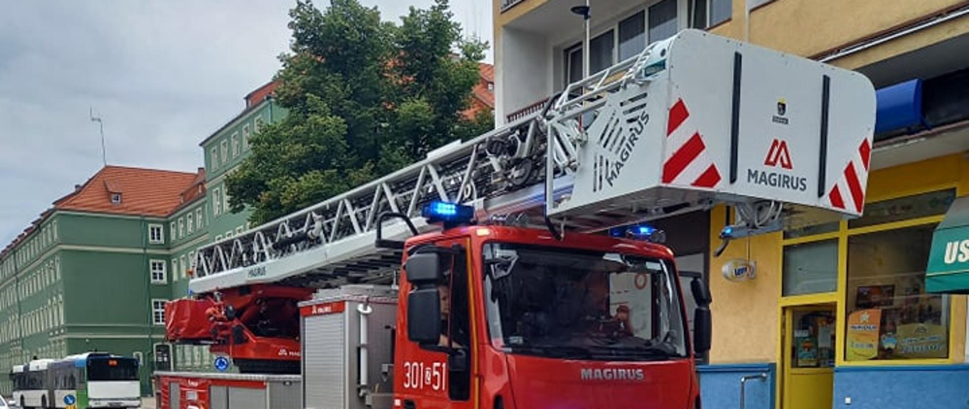 Samochód strażacki z drabiną mechaniczną, w tle budynki mieszkalne wielorodzinne oraz autobus komunikacji miejskiej.